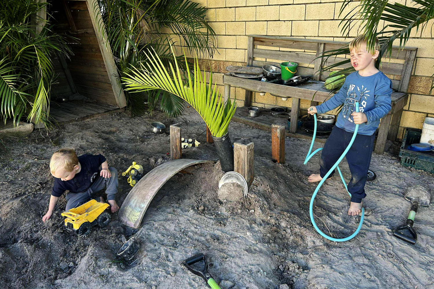 A photograph of two young children playing in an outdoor sandy area. They have built a makeshift construction site using various timber pieces. There are two small shovels on the ground. In the middle of the site is a black pipe sticking out of the sand holding a large green palm frond. One child is filling a yellow toy truck with sand and the other child is holding a length of hose pipe.