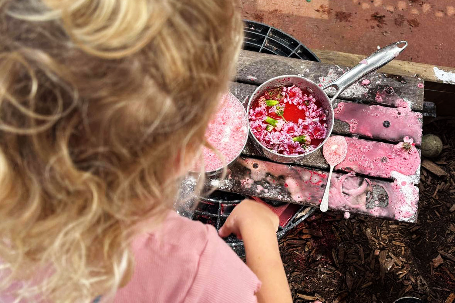 An overhead photograph of a child playing with pink liquid in a silver pot. Floating in the liquid are pink flower buds. The liquid has spilled over onto the wooden table.